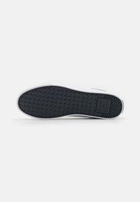 Semelle en caoutchouc noire d'une basket blanche avec une adhérence texturée et un marquage "Tommy Hilfiger" près du talon sur un fond uni.
