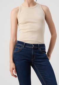 Beige tajt linne med rund halsringning och ärmlös design, parat med mörkblå jeans med orange sömmar och knappstängning.