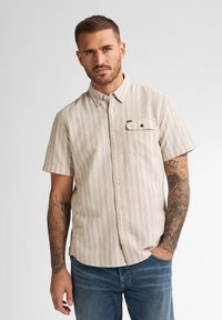 Camisa de manga corta, rayas beige con cuello abotonado, bolsillo frontal y textura de tela sutil. Combinada con jeans de mezclilla azul.