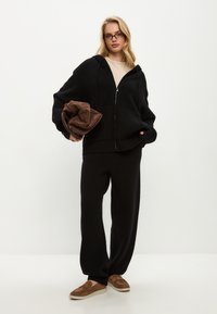 Černá mikina na zip s žebrovaným lemem, spárovaná s odpovídajícími širokými kalhotami. Drží hnědý, jemně strukturovaný pléd. Hnědé nazouvací boty.