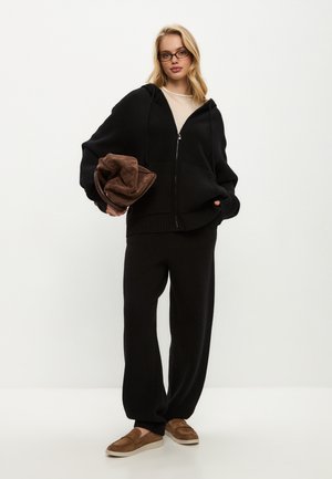 Felpa nera con zip e orlo a costine, abbinata a pantaloni larghi coordinati. Tieni in mano una coperta marrone a trama morbida. Scarpe marroni senza lacci.