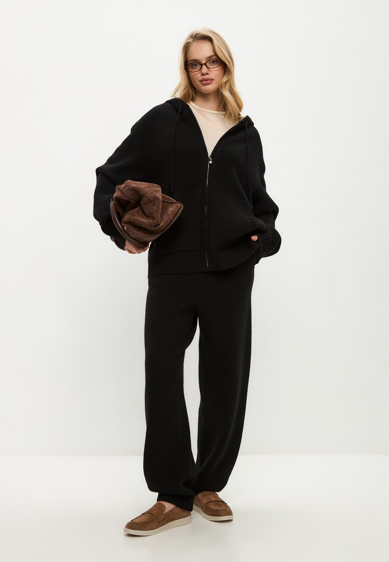 Černá mikina na zip s žebrovaným lemem, spárovaná s odpovídajícími širokými kalhotami. Drží hnědý, jemně strukturovaný pléd. Hnědé nazouvací boty.