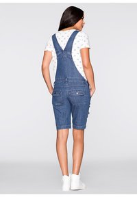 Salopette en denim, style court, couleur bleue. Dotée de bretelles réglables, d'une poche avant et de deux poches arrière. Portée sur une chemise blanche à motif cœur.