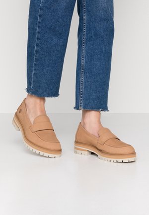 Persona che indossa mocassini beige in suede con suole bianche spesse e jeans blu a vita corta, su uno sfondo chiaro e semplice.