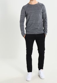 Man som står med händerna i fickorna, iförd en grå långärmad tröja, svarta byxor och vita sneakers på en enfärgad bakgrund.