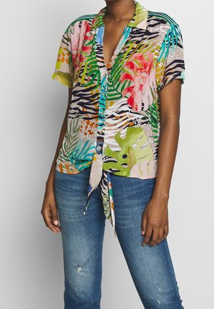 Nainen, jolla on värikäs, lyhythihainen, napitettava, trooppinen printtipaita, solmittu vyötärölle, ja siniset haalistuneet farkut.