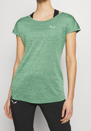 Frau trägt ein grünes, kurzärmeliges Sportshirt über einem schwarzen Sport-BH und schwarze Hose mit weißen Logos auf Shirt und Hose.