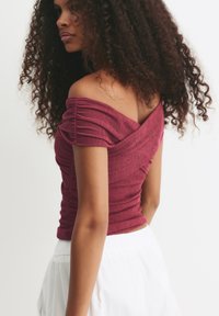 Femme aux cheveux bouclés portant un haut bordeaux texturé à épaules dénudées et une jupe blanche, tournée légèrement sur le côté devant un fond blanc uni.