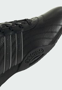 Black leather athletic shoe with three textured stripes, a rounded toe, and a rubber sole featuring the Adidas logo. Classic design elements evident.