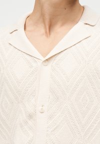 Πουλόβερ πλεκτό κρεμ χρώματος με γιακά και δύο κουμπιά, φορεμένο από ένα άτομο, που δείχνει λεπτομερή υφή και σχέδια στο ύφασμα.