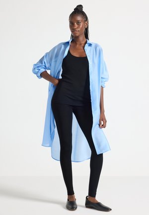 Femme aux cheveux tressés portant une chemise ample bleu clair à manches longues sur un débardeur noir, des leggings noirs et des chaussures noires sans lacets, debout.