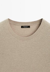 Geribbelde beige trui met een ronde halslijn en een merklabel, gemaakt van lichtgewicht, gestructureerde stof. Eenvoudig ontwerp zonder patronen.