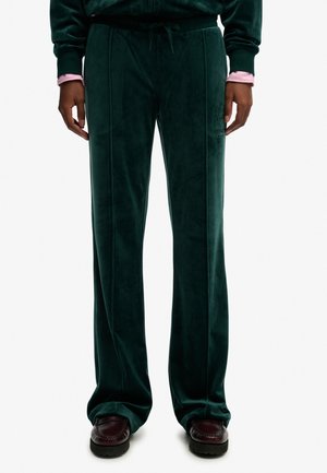 Ciemnozielone welurowe spodnie o szerokich nogawkach z przednimi szwami, noszone z ciemnobrązowymi butami i ciemnozielonym topem z różowymi mankietami.