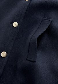 Marineblauwe stoffen jas met goudkleurige knopen, een strak ontwerp en een zijzak met een gestikte afwerking. Over het geheel een gladde textuur.