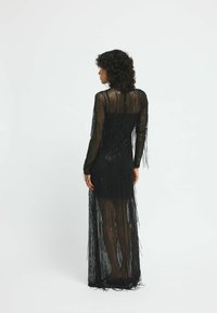 Rochie neagră cu franjuri, cu mâneci lungi, material transparent și o siluetă ajustată. Prezintă detalii verticale cu franjuri și un design până la podea.