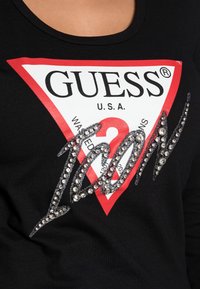 Czarna koszulka z grafiką w czerwono-białe trójkąty z napisem "GUESS® U.S.A." i "ICON" wyszytym kryształkami, podkreślająca odważny, nowoczesny design.