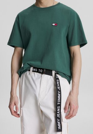 Mężczyzna w zielonej koszulce Tommy Jeans wpuszczonej w białe spodnie z luźno opuszczonym czarnym paskiem Tommy Jeans.