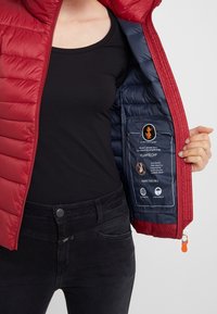 Rode donsjassen met een ritssluiting aan de voorkant, met een marineblauwe binnenkant en labels die materiaaleigenschappen beschrijven. Zwart tanktop en donkere spijkerbroek zijn zichtbaar.