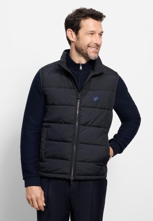 Uomo sorridente che indossa un gilet trapuntato nero sopra un maglione blu navy, in piedi con una mano in tasca, guardando di lato contro uno sfondo bianco.