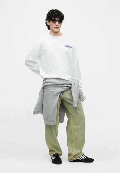 Jauns vīrietis ar cirtainiem matiem, valkājot melnas saulesbrilles, baltu krekla ar garām piedurknēm, brīvi pieguļošas dzelteni zaļas bikses, pelēku sporta krekla, sasietu ap vidukli, un melnus sporta apavus.