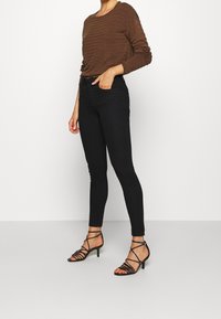 Donna con un top marrone a maniche lunghe, jeans skinny neri a vita alta e sandali neri con tacco a kitten su sfondo bianco semplice.