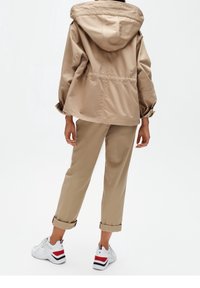 Persona con pantaloni beige arrotolati, giacca con cappuccio beige e sneakers bianche con strisce rosse e blu scuro, in piedi su uno sfondo bianco.
