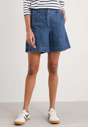 Denim shorts in dark wash with a relaxed fit, two front pockets, and visible stitching. Paired with white sneakers featuring green accents.