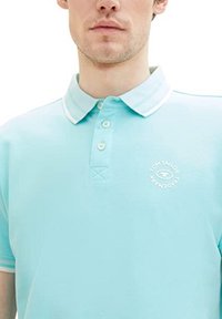 Polo bleu clair avec des finitions blanches au col. Il dispose d'une patte de boutonnage à trois boutons et d'un logo circulaire brodé sur la poitrine.