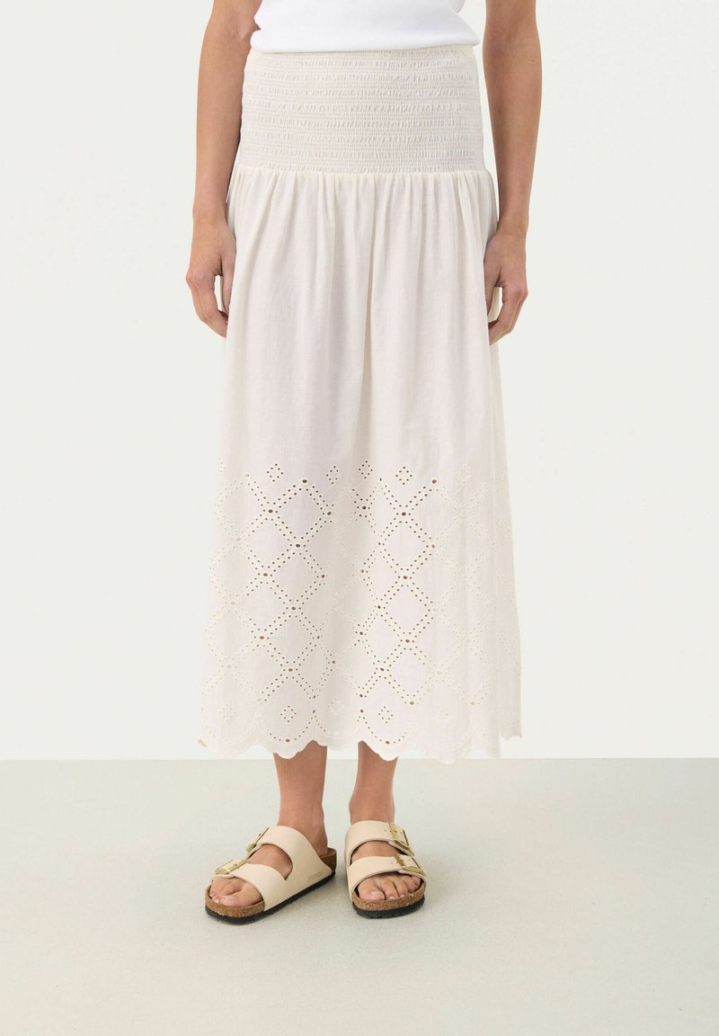 Lichtbeige katoenen rok met een smocked taille en een geborduurd uitgesneden patroon langs de zoom, gecombineerd met lichte sandalen.