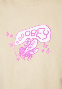 Beige bomullströja med en rosa grafik av en fluga och ordet "OBEY" i en pratbubbla, accentuerad med små stjärnor. Enkel design.