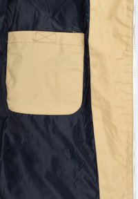 Poche en tissu beige cousue sur la doublure bleu foncé de la veste, à côté de la couture du zip beige sur le côté droit.