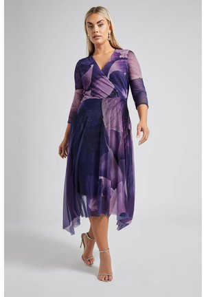 Lila Wickelkleid mit drei Viertelärmeln, floralem Muster, fließendem Saum und transparenter Überlage. Kombiniert mit silbernen Riemchen-Heels.