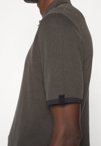 Homme portant un polo à manches courtes, gris foncé, en maille texturée avec une petite étiquette noire de logo sur l'ourlet de la manche gauche.