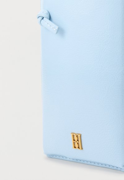 Γωνία πορτοφολιού από ανοιχτό μπλε δέρμα με αρχικά σε χρυσό ανάγλυφο και λεπτομέρεια από ραμμένη ετικέτα σε λευκό φόντο.