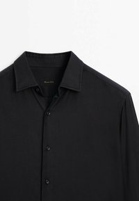 Chemise noire à manches longues, boutonnée avec un col classique, soigneusement repassée, présentée sur un fond blanc uni.