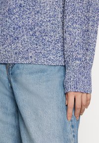 Sweter w kolorze niebieskim i fioletowym, dziergany, z teksturowanym wzorem; dziergane mankiety; noszony na jasnoniebieskich dżinsach.
