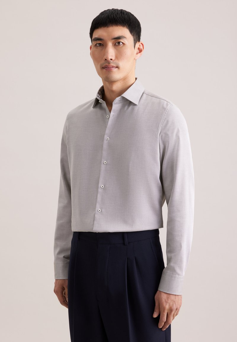 Homme debout portant une chemise légère gris clair boutonnée et un pantalon plissé bleu marine foncé sur un fond uni.