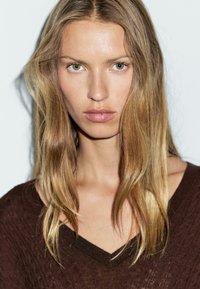 Nærbillede af en person med langt, lige, lyst brunt hår iført en mørkebrun, textured v-hals sweater, sat mod en ensfarvet baggrund.