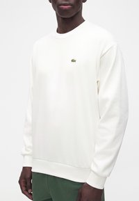 Homme portant un sweat-shirt blanc uni à manches longues avec un petit logo crocodile vert sur la poitrine et un pantalon vert foncé.