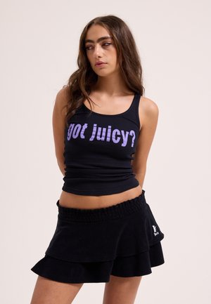 Młoda kobieta z długimi włosami, ubrana w czarny top na ramiączkach z napisem "got juicy?" oraz warstwową, czarną mini spódniczkę, na jednolitym tle.