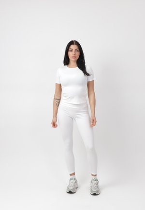 Frau mit langen dunklen Haaren, die ein weißes Kurzarmoberteil, weiße Leggings und weiße Turnschuhe trägt und vor einem schlichten Hintergrund steht.