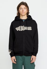 Haut zippé noir avec cordon de serrage, orné d'un grand logo graphique beige et d'un détail brodé blanc sur la manche gauche. Poches avant incluses.