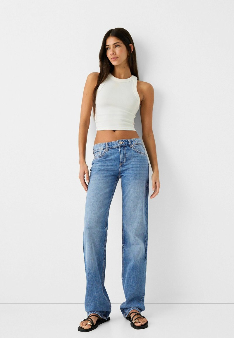 Top corto blanco acanalado, jeans de pierna ancha azul claro con lavado desgastado, que presentan un cierre de botón y cremallera, complementados con sandalias planas negras.