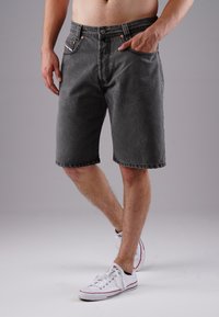Man wearing faded black denim shorts and white low-top sneakers, standing with one hand in pocket against a plain background.