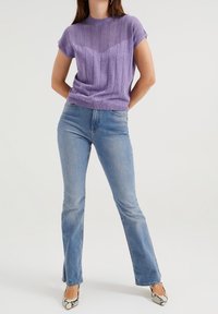 Kvinna som bär en lila kortärmad stickad tröja, ljusblå raka jeans med slits i framkant, och klackar i ormskinn med spetsig tå.