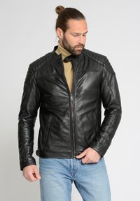 Sort skindmotorcykeljakke med polstrede skuldre, lynlåslukning og to sidelommer, båret over en beige turtleneck og blå jeans.