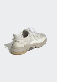 Baskets beiges en maille texturée et matériaux en caoutchouc, avec une semelle en forme de vague, des lacets blancs et une languette sur le talon.