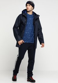 Navy waterproof jacket with a zip and snap closures, worn over a patterned blue shirt, paired with dark pants featuring a gray stripe, and black hiking boots.