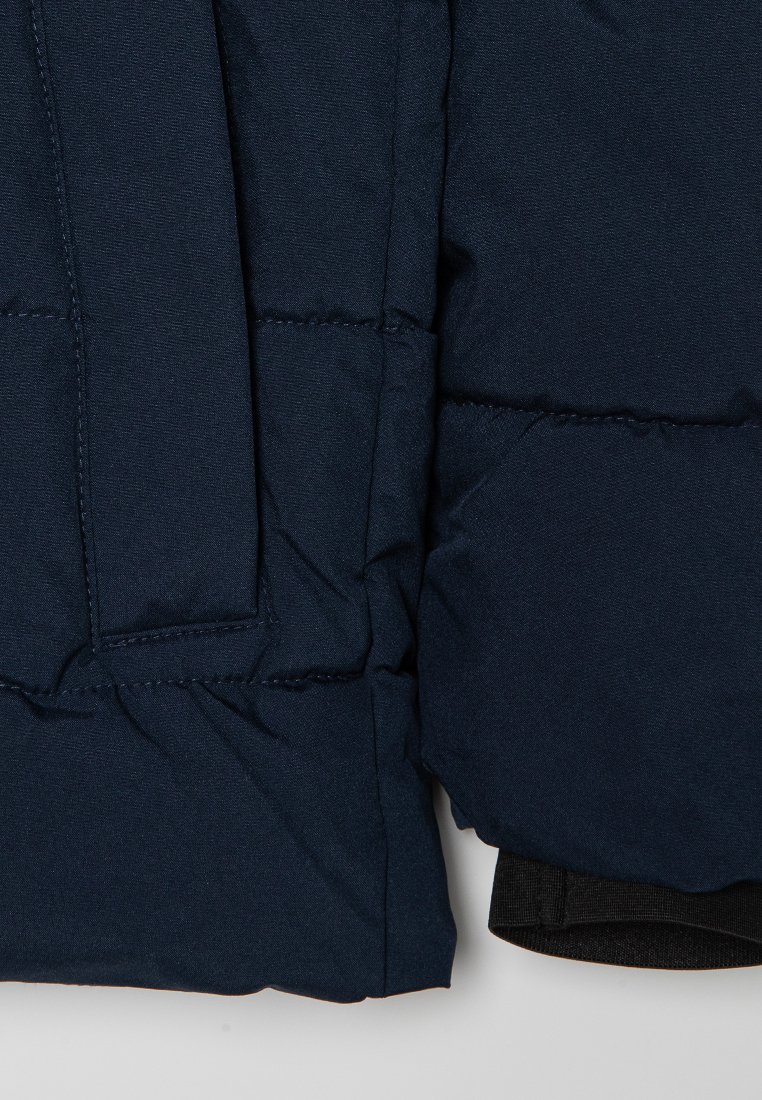 Chaqueta acolchada azul marino con textura de acolchamiento, presenta un bolsillo lateral y un dobladillo elástico negro. El material parece suave y duradero.