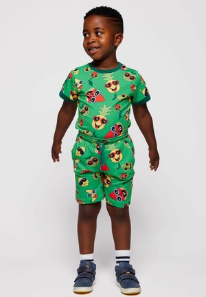 Ung dreng iført matchende grønne shorts og t-shirt med farverige frugtansigter iført solbriller, marineblå sneakers og hvide sokker.